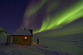 Man in front of Cape Swainson's cabin and northern lights. North East coast of Greenland