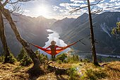 Woman with a sun hat sitting in an orange hammock, panoramic view of mountains with lake, Plansee, Tyrol, Austria, Europe