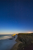 April 12, 2017 - The stars of Orion and Canis Major (including Sirius) setting head first into the west over the sea stack formations of the Twelve Apostles on the Great Ocean Road, Victoria, Australia, with the stacks beginning to be lit by light from the rising nearly full moon behind the camera in the east. The sky is blue with moonlight. 

