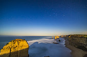 The Twelve Apostles sea stack formations on the Great Ocean Road, lit by the light of the rising nearly full moon off camera to the east, with Orion setting in the west over the cliffs. 
