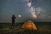 A person poses for a scene in Grasslands National Park, Saskatchewan, Canada, of stargazing with binoculars under the Milky Way on a dark moonless night. Grasslands is perfect for stargazing as it is a Dark Sky Preserve and the horizon is vast and unobstructed. . . Mars is bright to the left and the galactic centre is to the south at right. The view is overlooking the Frenchman River Valley.