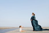 Side profile of a pregnant woman standing on the beach with her baby girl sitting in front of her