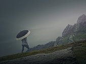 Rear view of a lonely man with umbrella stands on a rocky hill covered by haze. Moody and emotional scene with a lone stranger silhouette under the rain