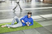 Businessman listening to music and reading book while relaxing on grass mat in an office lobby