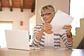 Woman at home office holding bills and looking at laptop
