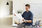 Smiling young man holding blueprint while sitting on desk at office