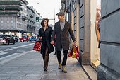 Smiling girlfriend and boyfriend walking with shopping bags on footpath in city
