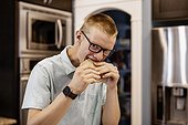 Young man standing in the kitchen at home and eating a sandwich, looking at the camera; Edmonton, Alberta, Canada