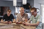 Father and two sons sit at the kitchen island at home using their smart phones, the father trying to talk to them; Edmonton, Alberta, Canada