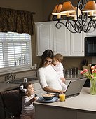 Caucasian mother holding baby  and typing on laptop computer with girl eating breakfast in kitchen.