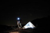 Stars illuminate the night sky as a man stands in front of his illuminated tent while wearing a headlamp in Palo Duro Canyon State Park.