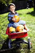 Young boy holds a pumpkin while seated in a red wagon filled with sqaush and pumpkin.