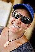 A woman wears sunglasses, visor and a seashell necklace while she smiles at the camera.