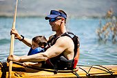 A man paddles a wooden kayak with a 3 year old boy in his lap.