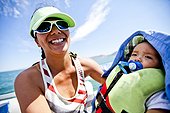 A Japanese-American women in her thirties smiles while wearing a seam foam green visor, while holding her 7 month old baby boy under the shade of a shirt while riding in a pontoon boat in the middle of Bear Lake, Utah.