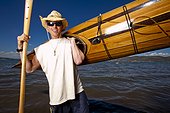 A man wears a straw hat as he poses for a portrait with a wooden kayak and paddle that he made while standing on the shore of Bear Lake, Utah.