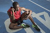 Male athlete sitting on all-weather running track and listening to music on smart phone, Barcelona, Spain
