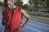 Male athlete standing on all-weather running track and listening to music on smart phone, Barcelona, Spain