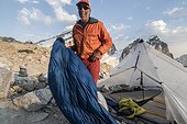 Smiling mountain climber standing near tent at mountain camp and holding sleeping bag, Bugaboo Mountains, British Columbia, Canada