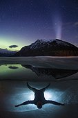 Man lying on ice on frozen Lake Minnewanka in winter under Aurora Borealis, Banff National Park, Alberta, Canada