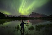 Aurora Borealis over photographer with tripod in lake and mountains in Banff National Park, Alberta, Canada