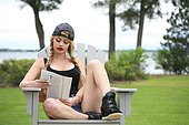 Beautiful young woman with braided hair reading magazine in her house backyard over looking the ocean, wearing baseball cap and shorts.