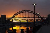 Newcastle's Tyne Bridge silhouetted by a dramatic sky that is reflected in the River Tyne. Streetlamps line the Quayside.