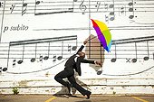 A man jumping and dancing with an umbrella outside. A young man jumping and dancing with an umbrella outside in front of a wall with musical notes on it.