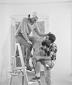 Young woman sitting on step ladder and man standing beside her while painting house