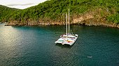 A man swims off the bow of a yacht anchored off shore from a dramatic cliff face.. Muskmelon Bay, Guana Island, British Virgin Islands.