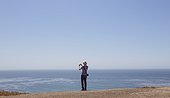 Man with shoulder bag stands by cliff's edge over the ocean and takes a photograph - wider shot.