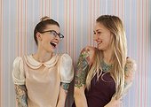 Two female friends with tattoos stand against a vintage wallpaper and laugh together as they look at each other.