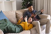 Diverse couple lying on couch and using tablet and smartphone in living room at home, copy space