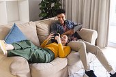 Happy diverse couple lying on couch, using tablet and smartphone in living room at home, copy space