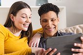 Happy diverse couple sitting on couch and using tablet in living room at home, copy space