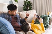 Diverse couple lying on couch and using smartphone in living room at home, copy space