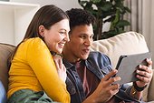 Happy diverse couple sitting on couch and using tablet in living room at home, copy space