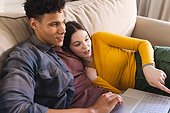 Happy diverse couple lying on couch and using laptop in living room at home, copy space