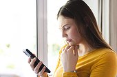 Focused caucasian woman using standing by window and using smartphone at home, copy space