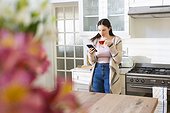 Focused caucasian woman standing, holding tea cup, using smartphone in kitchen at home, copy space