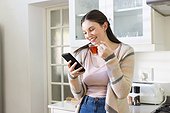Happy caucasian woman standing, holding tea cup, using smartphone in kitchen at home, copy space
