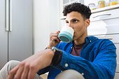 Concerned biracial man sitting on floor and drinking tea in kitchen at home, copy space