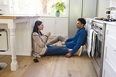Happy diverse couple sitting on floor, discussing, holding tea cups in kitchen at home, copy space
