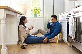 Happy diverse couple sitting on floor, discussing and drinking tea in kitchen at home, copy space