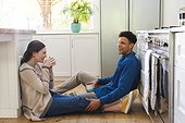 Happy diverse couple sitting on floor, discussing and drinking tea in kitchen at home, copy space