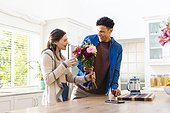 Happy diverse couple holding flowers and holding tea cup in kitchen at home, copy space