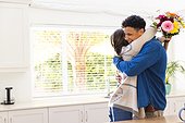 Happy diverse couple holding flowers and embracing in kitchen at home, copy space