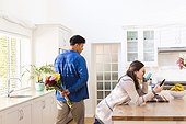 Diverse couple holding flowers behind back and using smartphone in kitchen at home, copy space