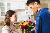 Happy diverse couple holding flowers and looking at each other in kitchen at home, copy space