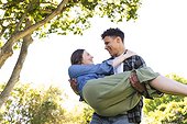 Happy diverse man holding woman in arms in air, looking at each other in sunny garden, copy space